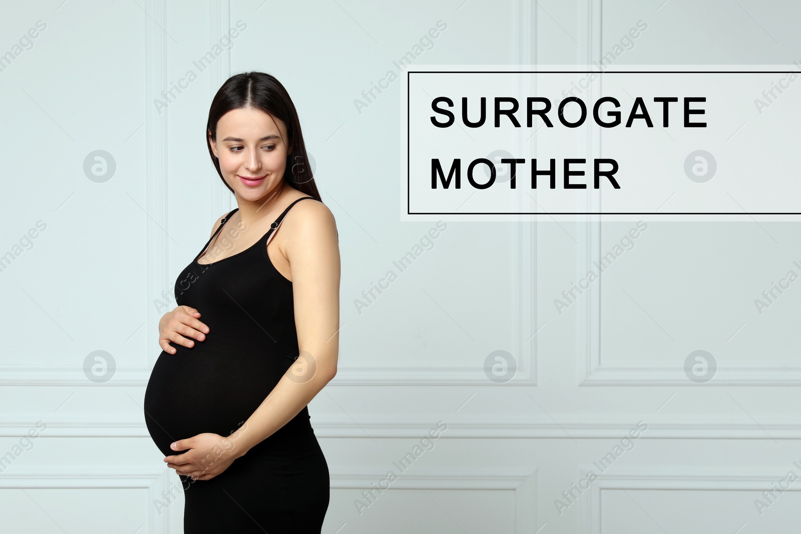 Image of Surrogate mother. Pregnant woman touching her belly near white wall indoors