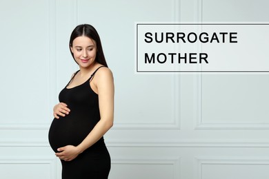 Surrogate mother. Pregnant woman touching her belly near white wall indoors