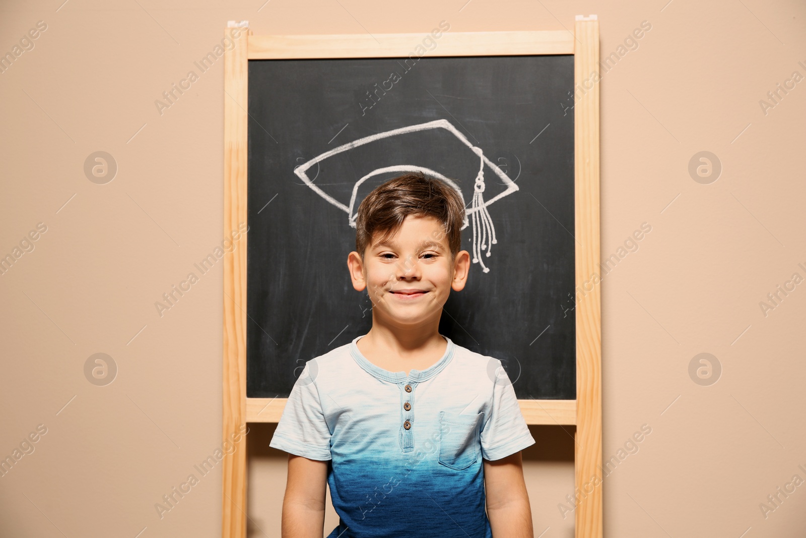 Photo of Cute little child standing at blackboard with chalk drawn academic cap. Education concept