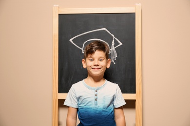Photo of Cute little child standing at blackboard with chalk drawn academic cap. Education concept