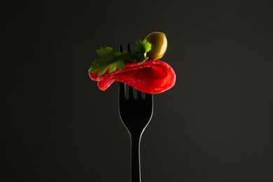 Photo of Fork with tasty slice of salami, olive and parsley on black background