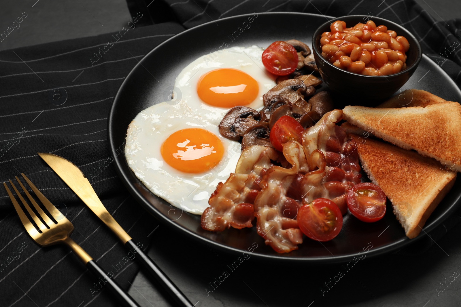 Photo of Plate with fried eggs, mushrooms, beans, bacon, tomatoes and toasted bread on black table, closeup. Traditional English breakfast