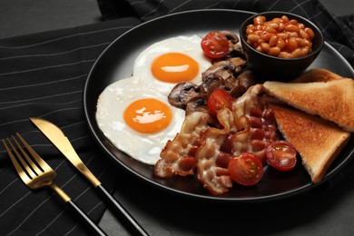 Photo of Plate with fried eggs, mushrooms, beans, bacon, tomatoes and toasted bread on black table, closeup. Traditional English breakfast