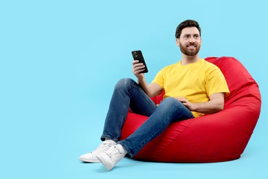 Photo of Smiling man with smartphone sitting on bean bag against light blue background. Space for text