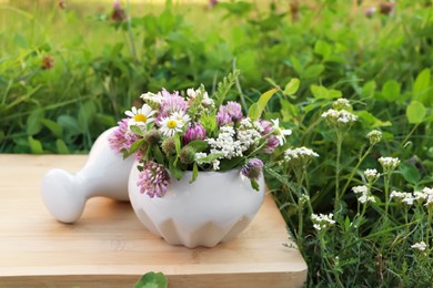 Photo of Ceramic mortar with pestle, different wildflowers and herbs on wooden board in meadow