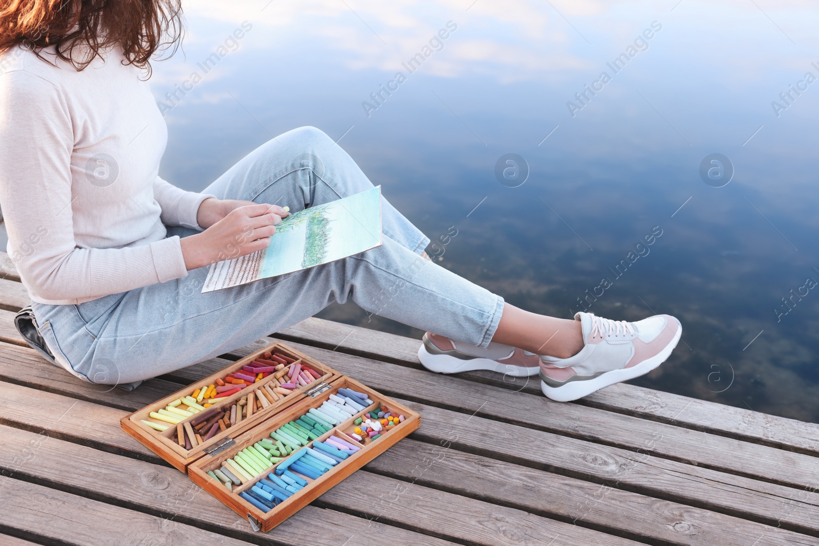 Photo of Woman drawing with soft pastels on wooden pier near river, closeup