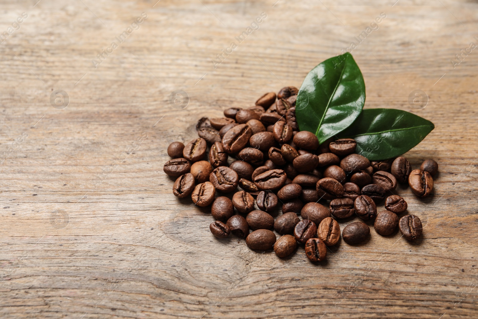 Photo of Pile of coffee beans and fresh green leaves on wooden table, space for text