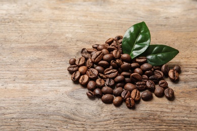 Photo of Pile of coffee beans and fresh green leaves on wooden table, space for text