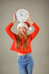 Young beautiful woman in Santa hat holding big clock on grey background. Christmas celebration