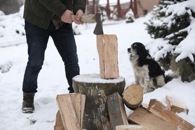 Photo of Man chopping wood with axe next to cute dog outdoors on winter day, closeup