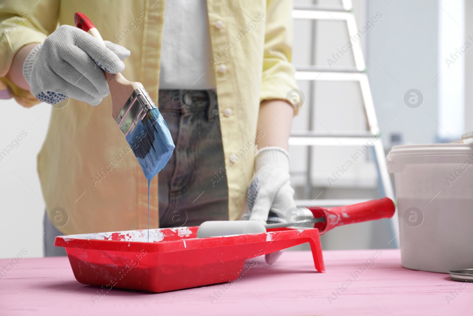 Photo of Woman taking light blue paint with brush from tray at pink wooden table indoors, closeup. Space for text