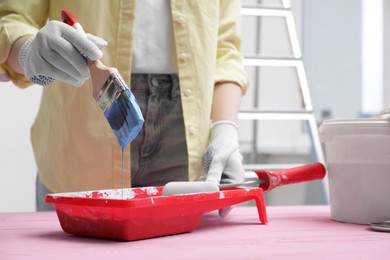 Woman taking light blue paint with brush from tray at pink wooden table indoors, closeup. Space for text