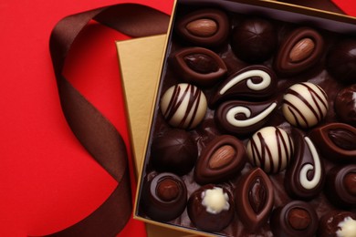 Box of delicious chocolate candies and brown ribbon on red background, flat lay