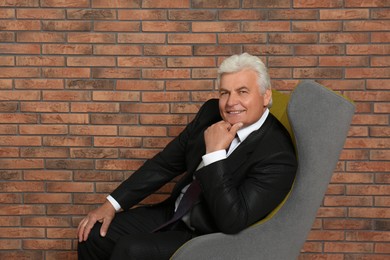 Photo of Happy mature businessman sitting in armchair near brick wall, space for text