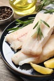Photo of Pieces of raw cod fish, rosemary and lemon on wooden table, closeup
