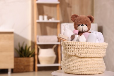 Photo of Knitted basket with baby cosmetic products, bath accessories and toy bear on white table indoors. Space for text