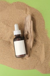 Photo of Bottle with serum and bark on sand against green background, top view. Cosmetic product