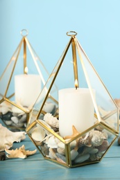 Stylish glass holders with burning candles, seashells and pebbles on light blue wooden table