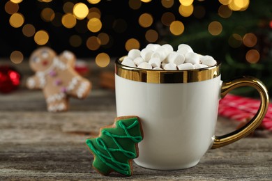 Delicious hot chocolate with marshmallows and Christmas tree gingerbread cookie on wooden table against blurred lights, closeup. Space for text