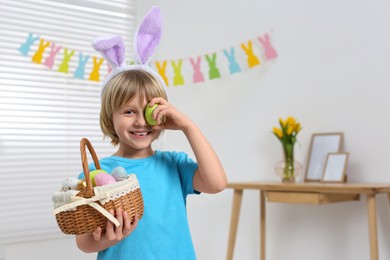 Happy boy in bunny ears headband holding wicker basket with painted Easter eggs indoors. Space for text