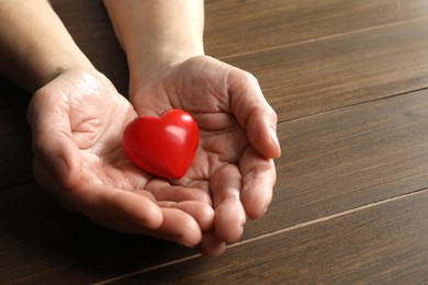 Photo of Elderly woman holding red heart in hands at wooden table, closeup. Space for text