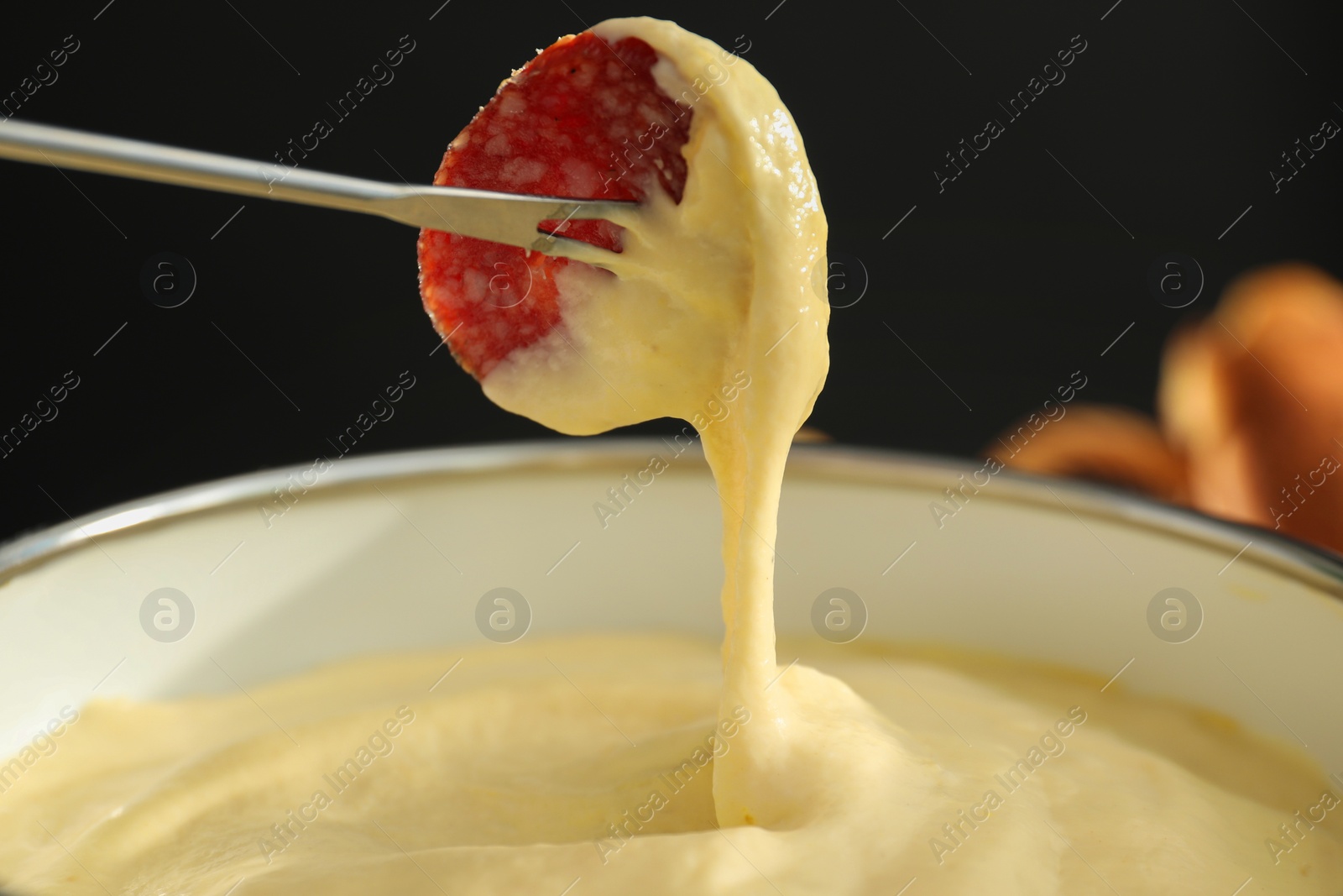 Photo of Dipping piece of sausage into fondue pot with melted cheese on black background, closeup