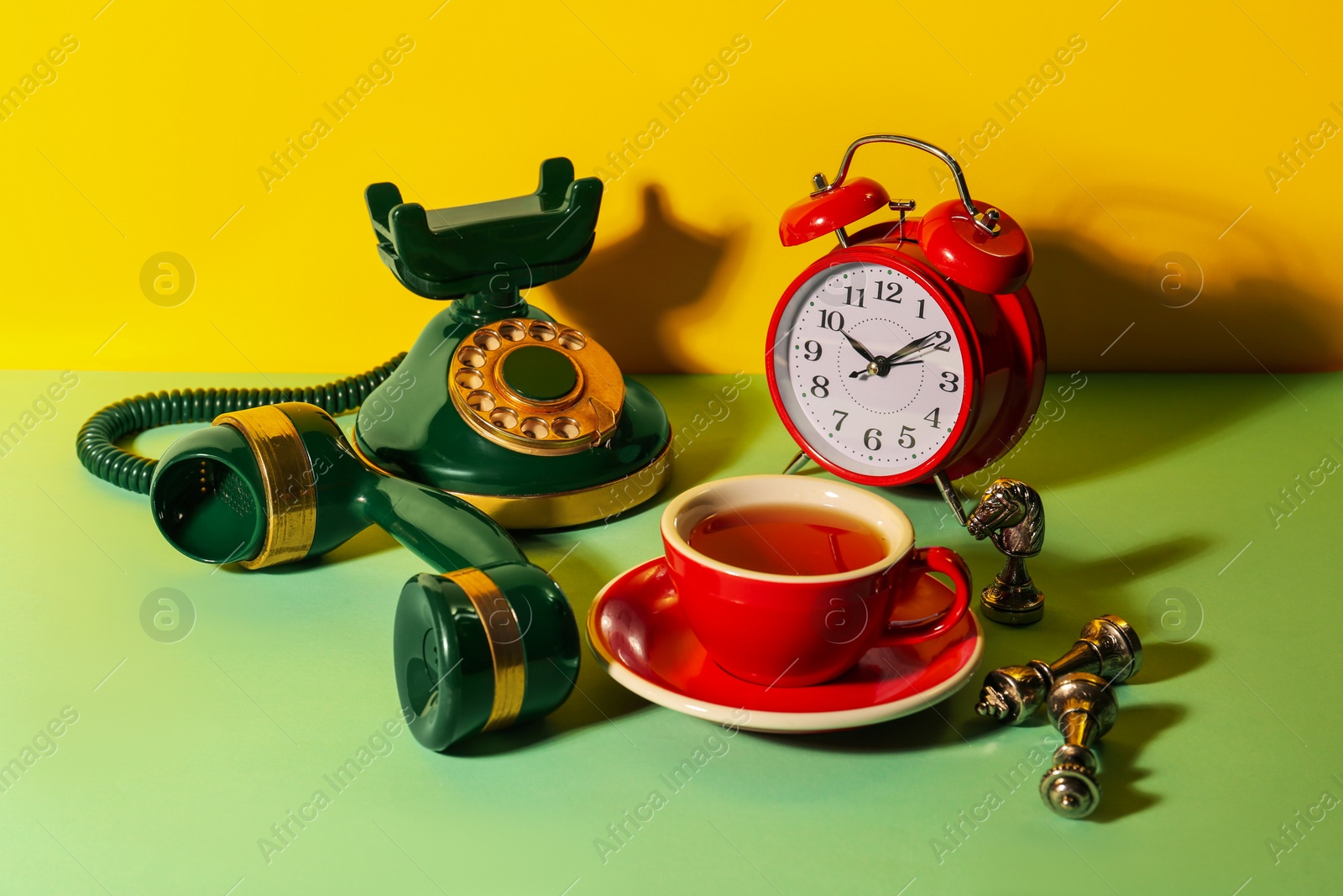 Photo of Red cup of tea, alarm clock, vintage telephone and chessmen on light green table against yellow background