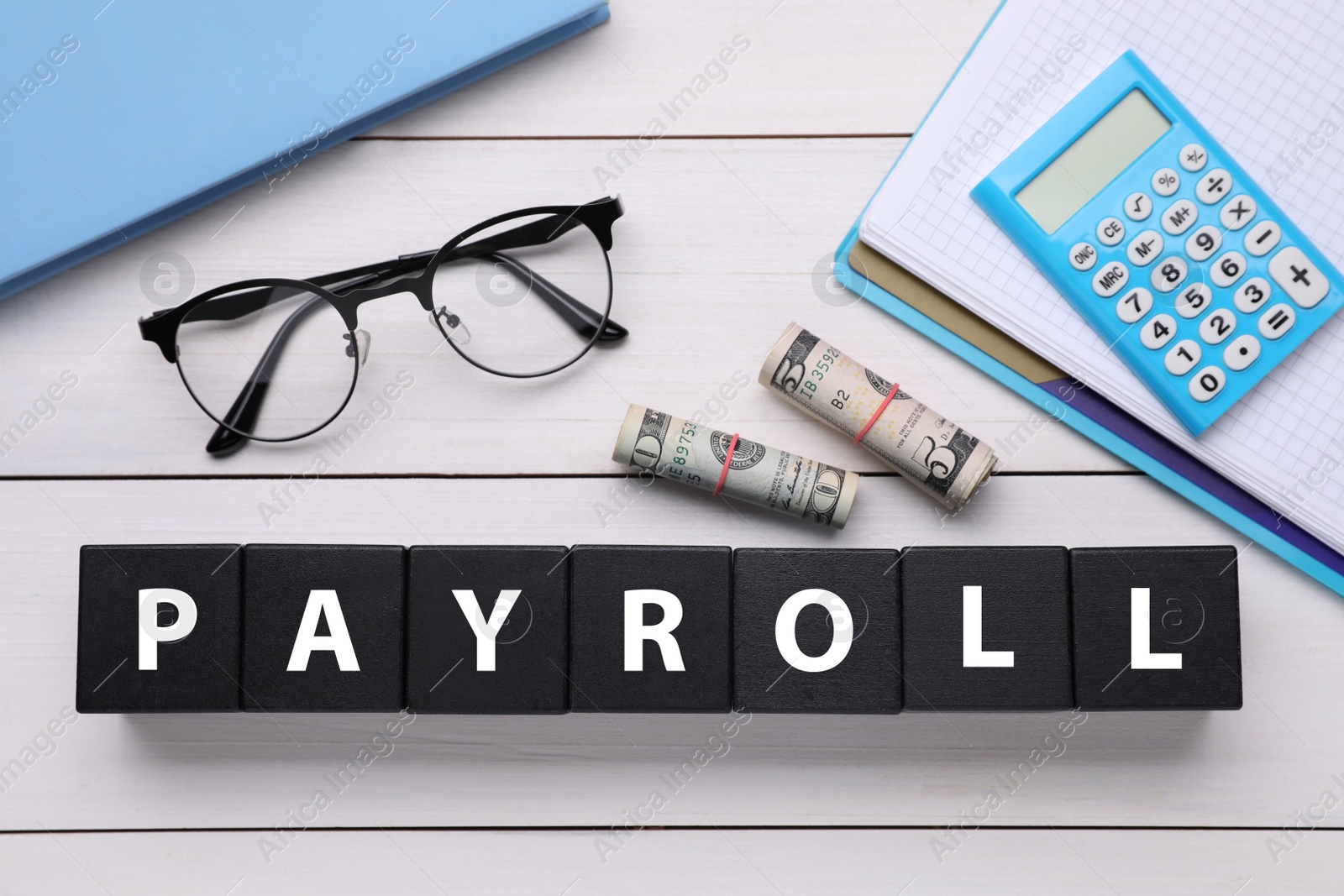 Photo of Word Payroll made of black cubes with letters, dollars, eyeglasses, notebooks and calculator on white wooden table, flat lay