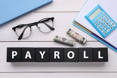 Word Payroll made of black cubes with letters, dollars, eyeglasses, notebooks and calculator on white wooden table, flat lay