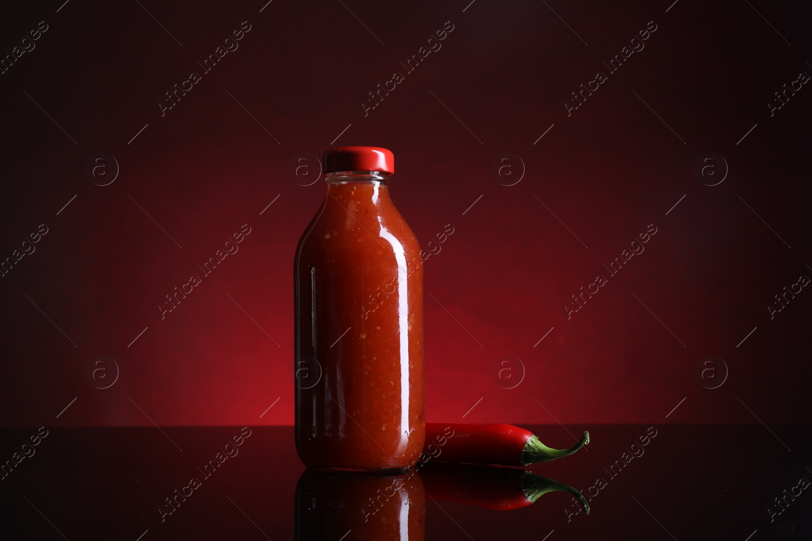 Photo of Spicy chili sauce in bottle and pepper against dark background