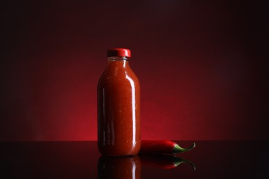 Photo of Spicy chili sauce in bottle and pepper against dark background