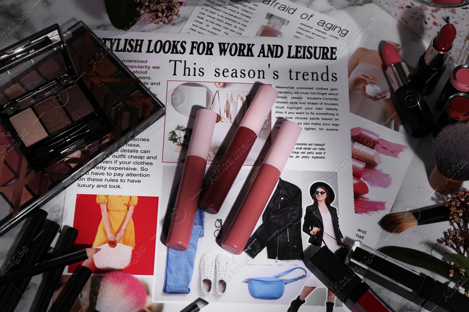 Photo of Bright lip glosses among different cosmetic products and fashion magazine on table, flat lay