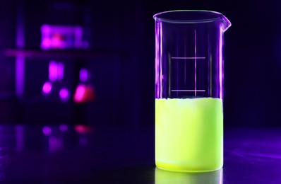 Photo of Laboratory beaker with luminous liquid on table against dark background, space for text