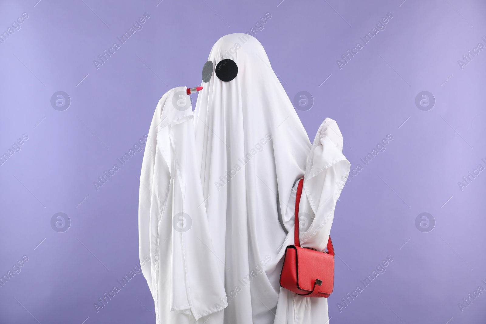 Photo of Glamorous ghost. Woman in white sheet with stylish bag and lipstick on violet background