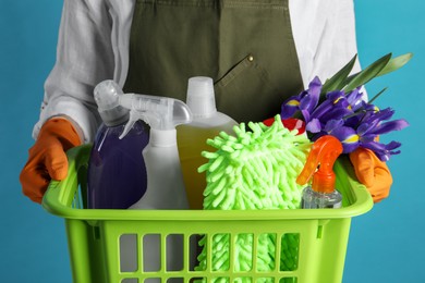 Spring cleaning. Woman holding basket with detergents, flowers and tools on light blue background, closeup