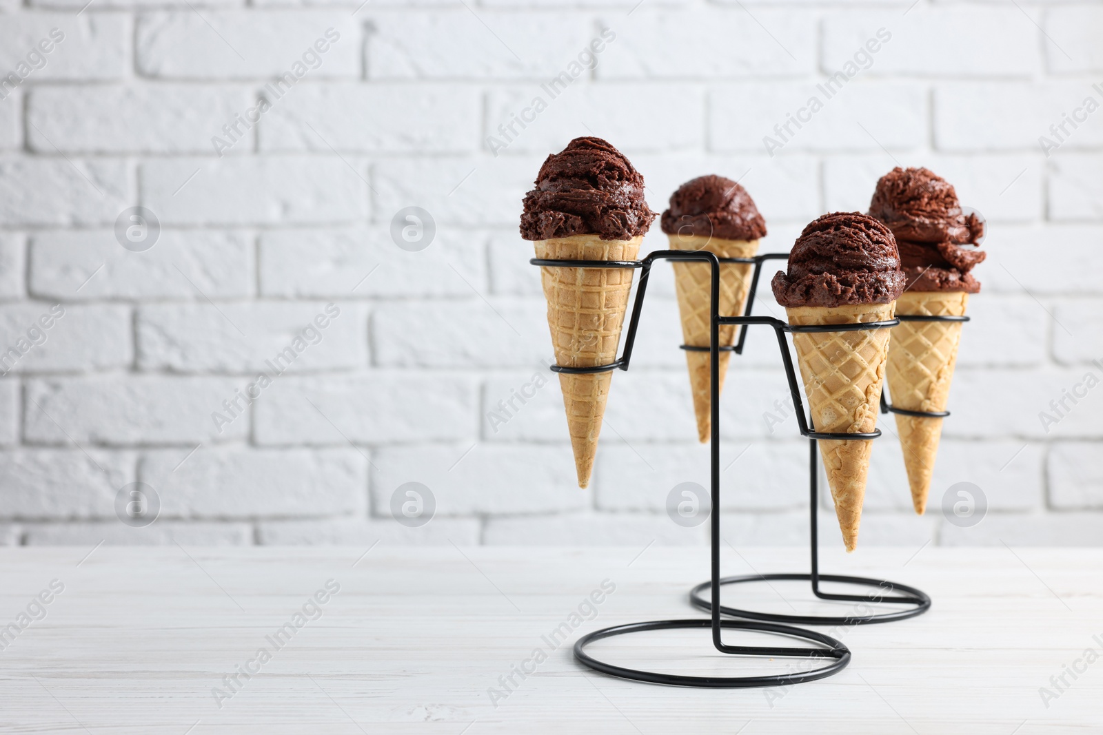 Photo of Chocolate ice cream scoops in wafer cones on white wooden table against brick wall, closeup. Space for text