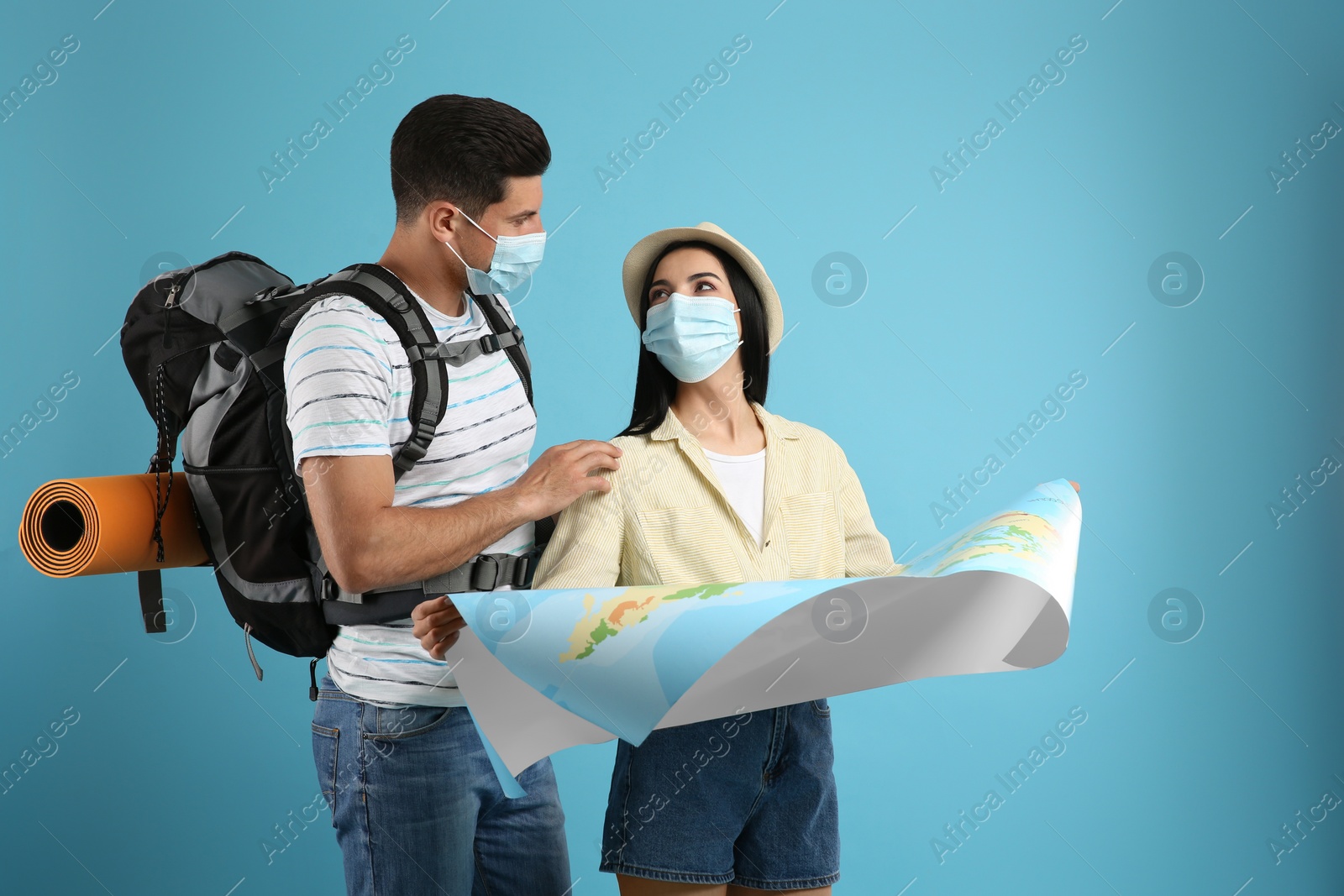 Photo of Couple of tourists in medical masks with map on light blue background. Travelling during coronavirus pandemic