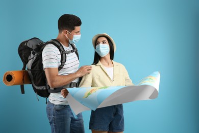 Photo of Couple of tourists in medical masks with map on light blue background. Travelling during coronavirus pandemic