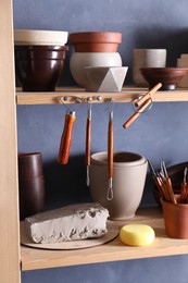 Photo of Set of different crafting tools and clay dishes on wooden rack in workshop, closeup