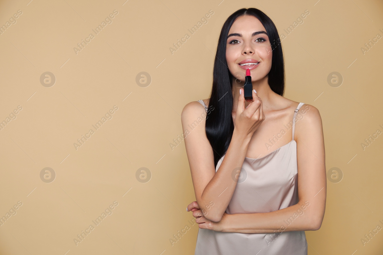 Photo of Beautiful young woman applying nude lipstick on beige background, space for text