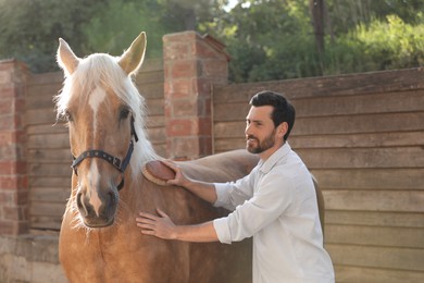 Photo of Man brushing adorable horse outdoors. Pet care