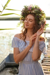 Photo of Young woman wearing wreath made of beautiful flowers near river on sunny day