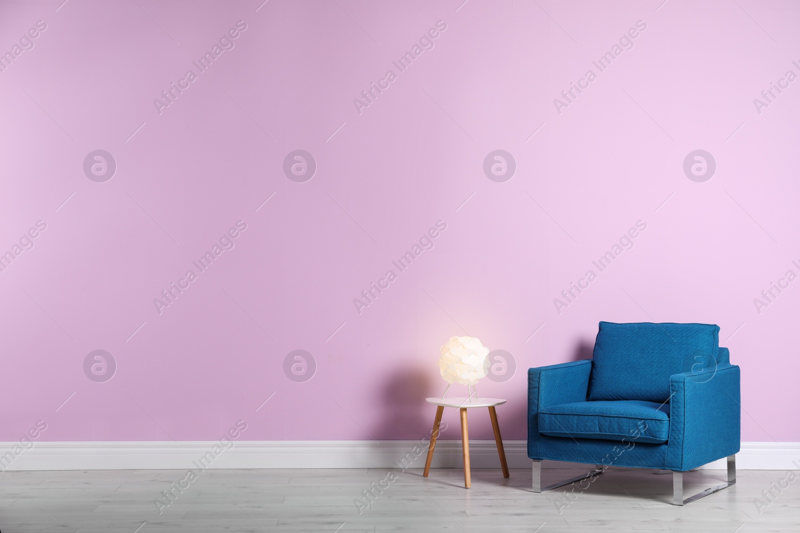 Photo of Comfortable armchair and lamp on table near color wall with space for text. Interior element