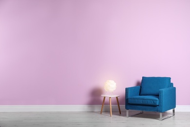 Comfortable armchair and lamp on table near color wall with space for text. Interior element