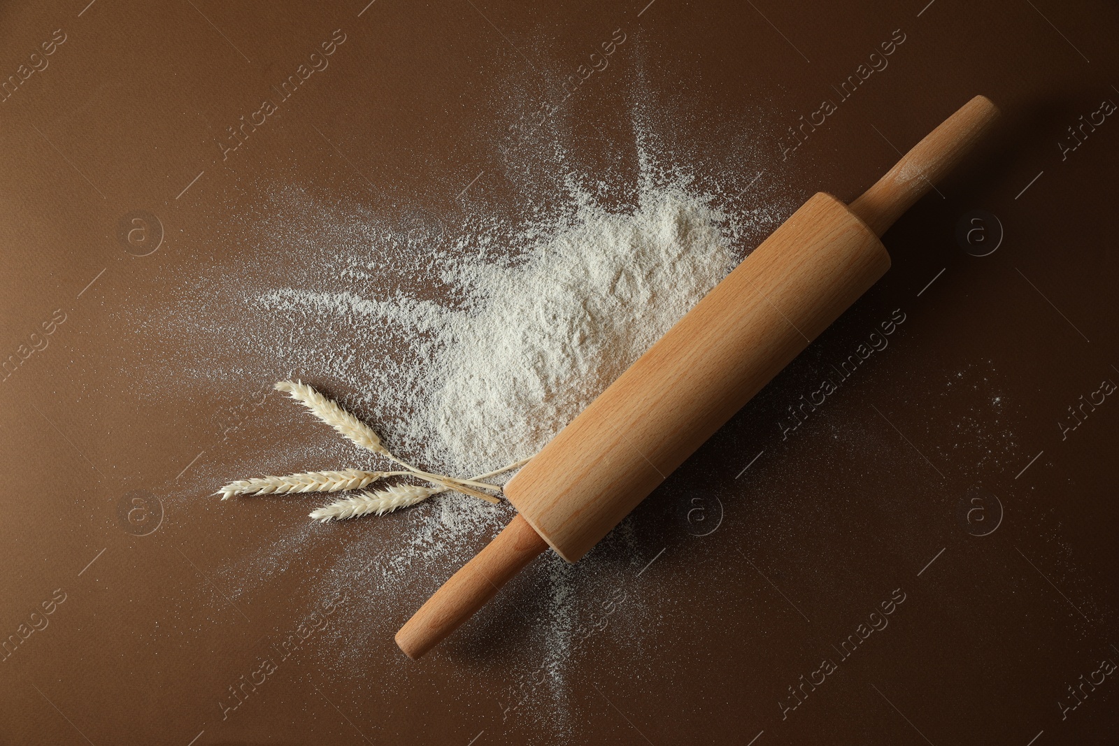 Photo of Flour, spikelets and rolling pin on brown table, top view