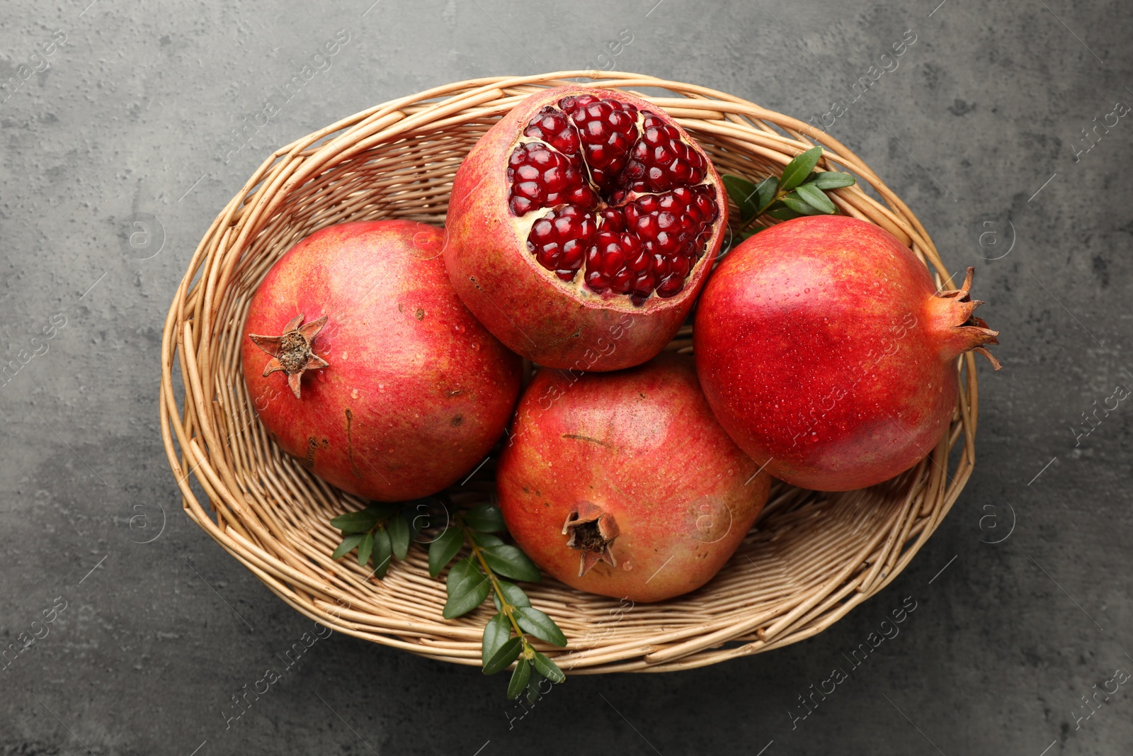 Photo of Fresh pomegranates and green leaves in wicker basket on grey table, top view