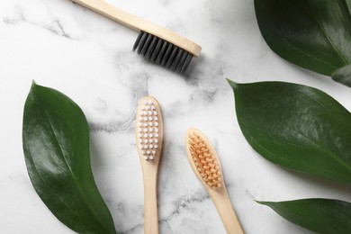 Bamboo toothbrushes and green leaves on white marble table, flat lay