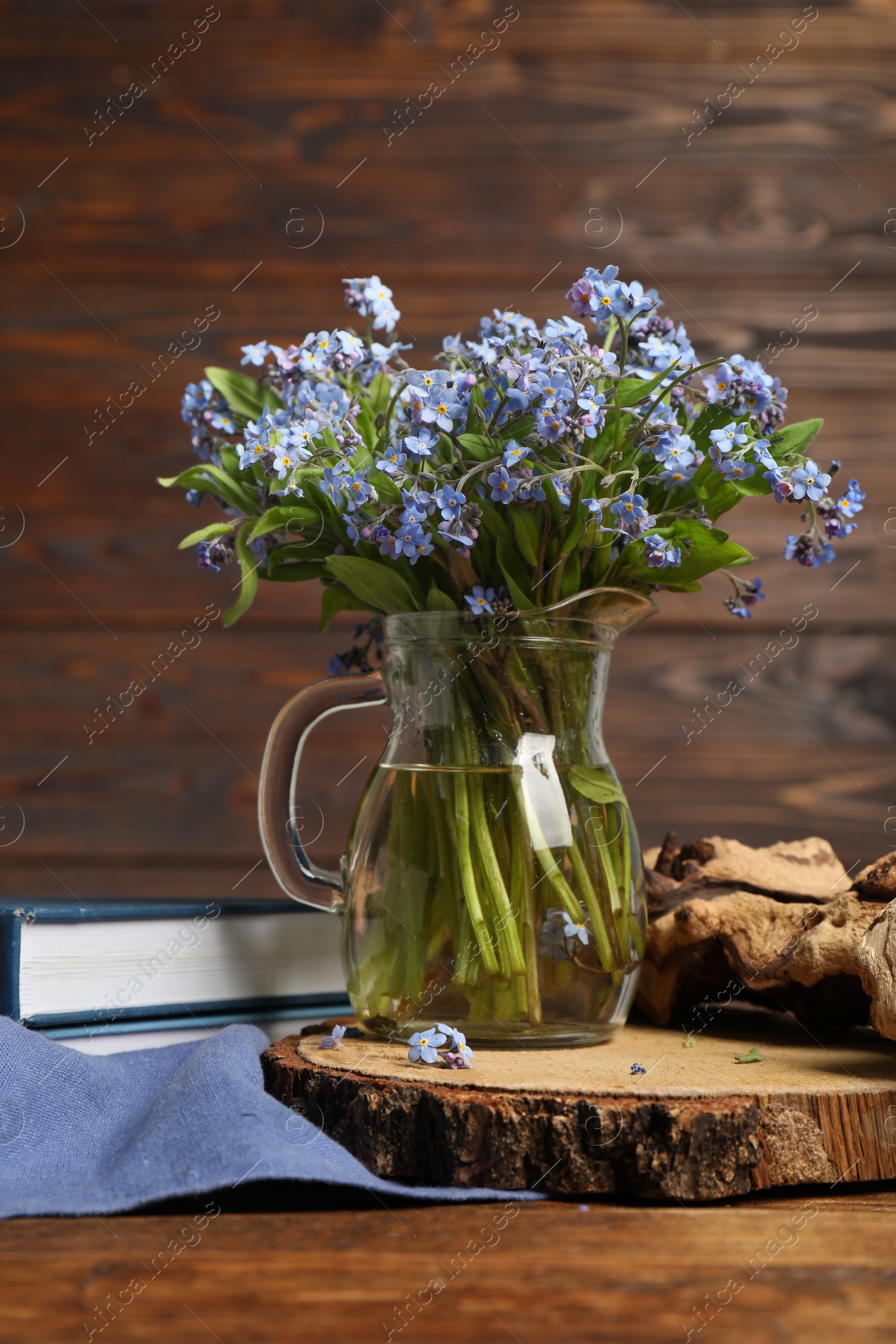 Photo of Bouquet of beautiful forget-me-not flowers in glass jug and book on wooden table