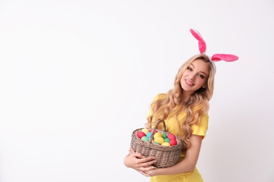 Photo of Beautiful young woman in bunny ears headband holding basket with Easter eggs on light background, space for text
