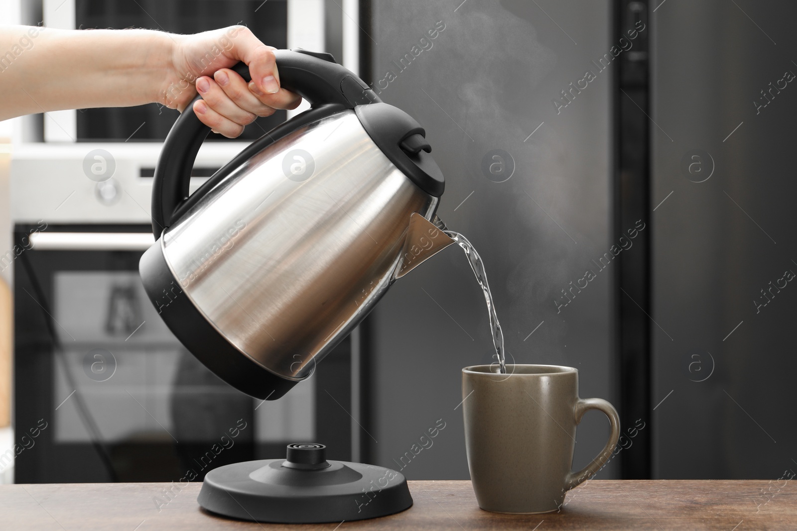 Photo of Woman pouring hot water from electric kettle into cup in kitchen, closeup